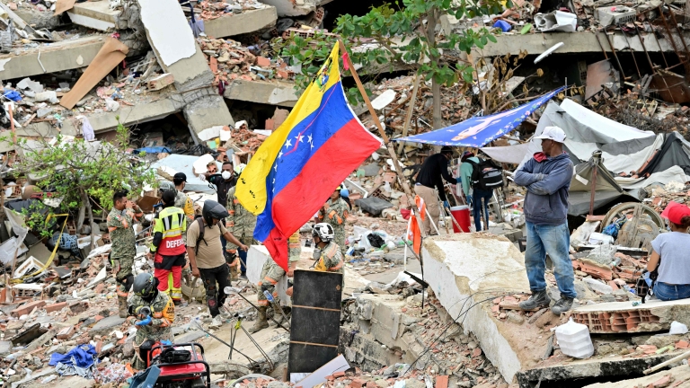 Members of a Mexican Army rescue unit and volunteers work amid the rubble of collapsed buildings as a Venezuelan flag stands in the foreground in Caraballeda, La Guaira state, Venezuela, on July 3, 2026. The death toll from Venezuela's twin earthq...